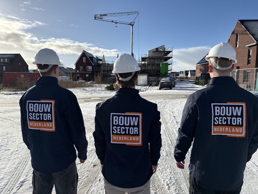 Drie vakmensen van Bouwsector Nederland met witte helm en werkjas kijken uit over een besneeuwde bouwplaats